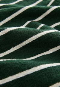 Zielony materiał z białymi ukośnymi paskami, o teksturowanej, miękkiej powierzchni. Wzór oferuje klasyczną kompozycję z kontrastującymi kolorami.