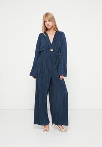 Navy zila plāna kombinezona ar garām piedurknēm un dziļu V-veida kakla izgriezumu. Iespējas ietver apaļu izgriezumu jostasvietā un platus, plūstošus kājstarpus.