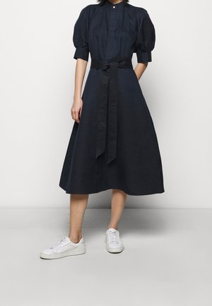 Kvinde iført en navy midi kjole med puffede ærmer og bredt bælte, kombineret med hvide sneakers, stående mod en ensfarvet baggrund.