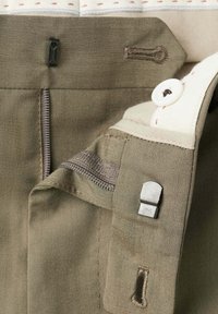 Primer plano de pantalones beige mostrando cremallera, cierre de gancho y barra, botón y ojales en el interior de la cintura.