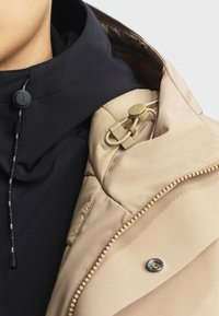 Beige buitenjasje met een zwarte binnenkraag. Kenmerkt zich door een trekkoordsluiting en een drukknoop. Soepele textuur met gestikte details.