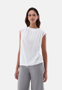 Blusa bianca senza maniche con collo plissettato, realizzata in tessuto leggero, abbinata a pantaloni larghi grigi. Texture morbida, design semplice.