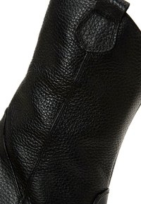 Bota de cuero negro con acabado de grano texturizado, caña alta y ligera curvatura en la parte superior. Presenta una costura en el lateral.