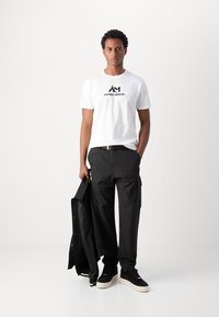 T-shirt blanche avec logo noir, pantalon cargo noir, veste noire tenue à la main et baskets noires avec semelles blanches. Éléments de design simples.