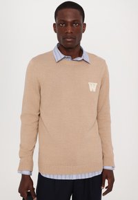 Beež kudumiga sall, millel on soonikust mansetid ja äär, millel on väike valge täht "W" logo. Kandmine helesinise triibulise kraega särgi peal.