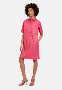 Rosa Kurzarmhemdkleid mit Knopfleiste, Kragen und Seitentaschen. Glattes, leichtes Material. Kombiniert mit rosa Absatzsandalen.
