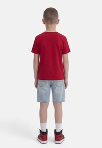 Child standing facing away wearing a red t-shirt, light denim shorts, white socks, and red and black sneakers against a white background.