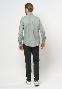 Maglietta verde chiaro a maniche lunghe con piega posteriore, orlo curvo e polsini con bottone, abbinata a pantaloni grigio scuro e sneaker bianche.