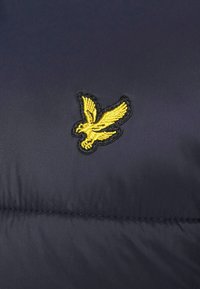 Granatowa kurtka puchowa z teksturowaną powierzchnią, widocznymi szwami i żółtym haftowanym logo orła na klatce piersiowej.