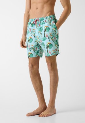 Mann trägt türkisfarbene Badehose mit Tukan- und tropischem Blätterdruck, steht barfuß mit einer Hand in der Tasche vor weißem Hintergrund.