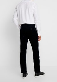 Tmavě modré kordové kalhoty ve rovné siluetě, doplněné bílou košilí s knoflíky. Černé tenisky dotvářejí outfit, pohled zezadu.