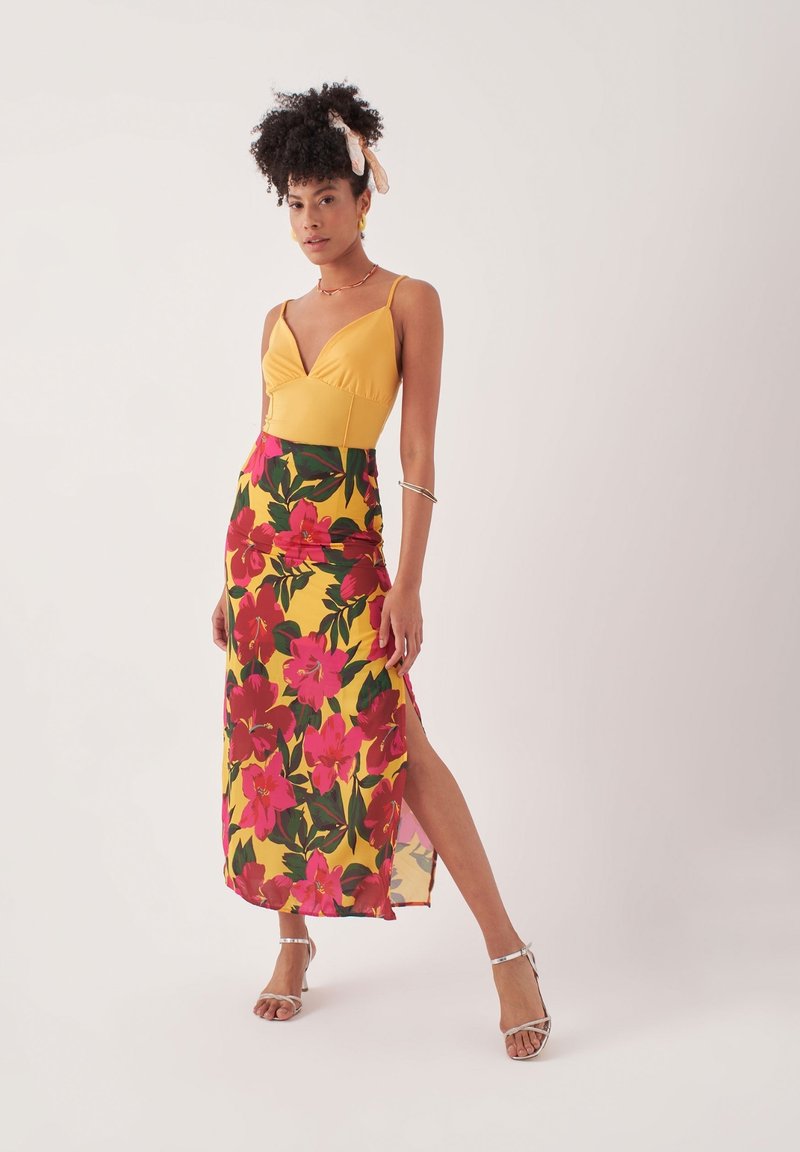 Geel aangesloten bovenstuk met dunne bandjes, in combinatie met een bloemenprint rok in roze en groen, met een hoge zijsplit. Zilveren sandaaltjes met banden.