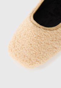 Beige fluffig toffel med rundad fyrkantig tå och svart inredning, vilande på en vit yta.