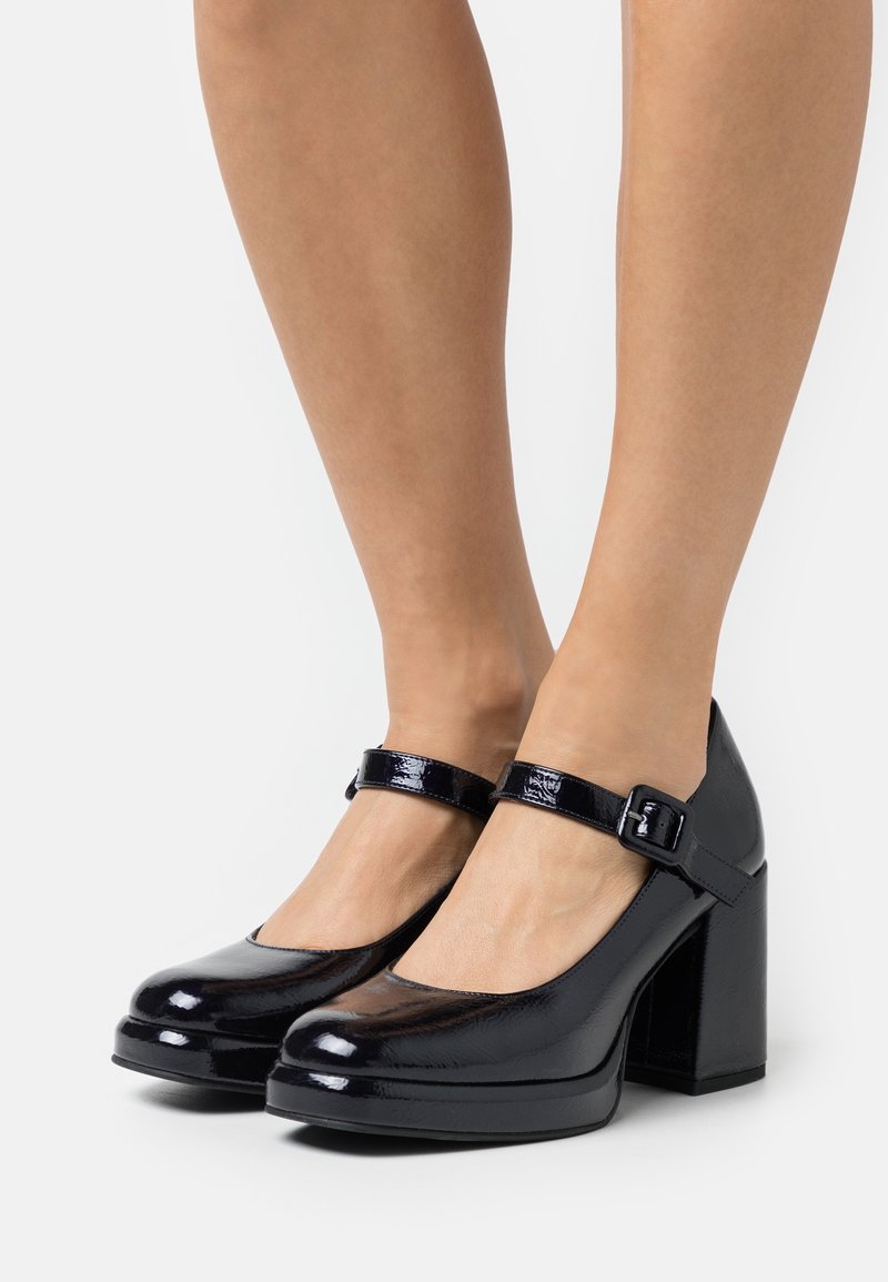 Escarpins Mary Jane noirs en cuir verni brillant avec brides chevilles et talons carrés blocs portés pieds nus sur un fond uni.