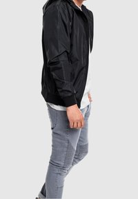 Black lightweight bomber jacket with a hood, front zipper, and ribbed cuffs, paired with light gray fitted pants featuring textured accents.