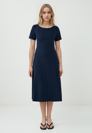 Modella che indossa un vestito blu navy aderente con maniche corte e orlo al ginocchio, in piedi su uno sfondo bianco. Infradito nere completano il look.
