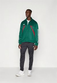 Grønn treningsjakke med glidelås og røde detaljer, hvite striper på ermene; kombinert med svarte treningsbukser med hvite striper og logo.