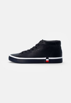 Czarny sneaker mid-top z białym paskiem na podeszwie, subtelnym podpisem Tommy Hilfiger z boku oraz małym czerwono-biało-niebieskim detalem flagi przy pięcie.