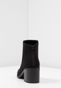 Stivaletto nero con tacco alto, realizzato in tessuto strutturato. Presenta un design a ritaglio geometrico e una zip esposta sul retro.