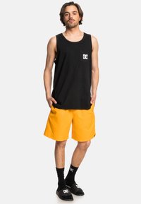 Schwarzes Baumwoll-Tanktop mit Tasche, lebendige gelbe Shorts, schwarze Socken mit weißem Logo und schwarze Sandalen mit Logodetails.