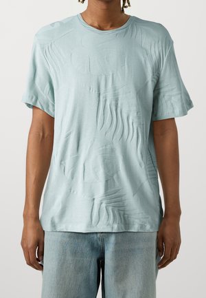 Homme portant un t-shirt à manches courtes bleu clair avec un motif texturé de feuilles tropicales et un jean denim bleu clair, sur fond uni.