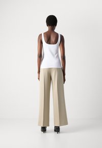 Vrouw die met de rug naar de camera staat, gekleed in een witte tanktop, beige wijde broek en zwarte hoge hakken, op een effen witte achtergrond.
