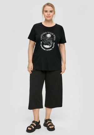 Schwarzes Baumwoll-T-Shirt mit weißem graphischem Text und floralem Design. Kombiniert mit schwarzen, weit geschnittenen Hosen und schwarzen Sandalen.