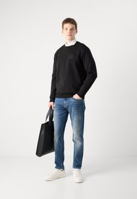 Black sweatshirt with a small square detail, blue jeans, white sneakers, and a black tote bag. Model stands against a plain background.