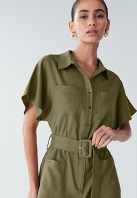 Kratek olivno zeleni oblek z ovratnikom, dvema žepoma na prsih in poudarjenim pasom z pravokotnim pasom. Gladek tekstura tkanine.