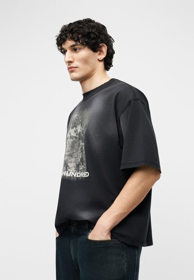 Άνδρας με σγουρά μαλλιά που φοράει μια φαρδιά μαύρη μπλούζα με σχέδιο και σκούρο τζιν, με τα χέρια στις τσέπες, κοιτάζοντας προς τα αριστερά με ουδέτερη έκφραση.