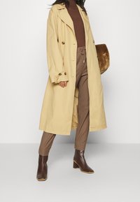 Trench-coat beige, col roulé côtelé marron, pantalon à carreaux et bottines à talons marron. Tissu doux avec design croisé à double boutonnage et détails de boutons.