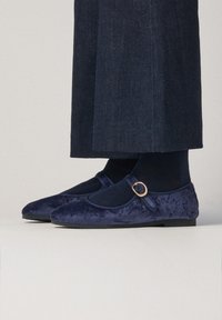 Flats Mary Jane de terciopelo azul marino con una correa y detalle de hebilla dorada. Punta redondeada y suela plana; combinados con pantalones de mezclilla oscuros.