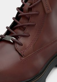 Close-up de uma bota de couro marrom com bico arredondado, costura detalhada, atacadores escuros, fecho de metal para os atacadores e sola preta.