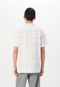 Camicia a maniche corte in tessuto bianco con superficie testurizzata, caratterizzata da un motivo a griglia nera. Con colletto e orlo dritto, indossata con pantaloni grigi.