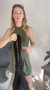 Vestido verde oliva com um decote alto e design peplum. Modelo segura uma alça preta texturizada, mostrando o seu padrão trançado. Fundo neutro.