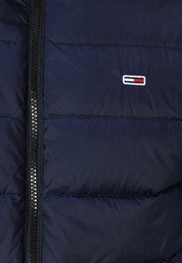 Jachetă puffer de culoare bleu marin, cu închidere prin fermoar și detalii cu cusături orizontale. Prezintă un logo brodat în alb, roșu și albastru.