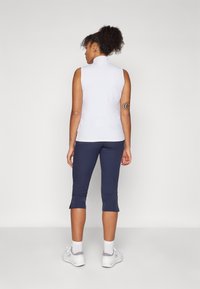 Top blanco sin mangas con cuello alto, combinado con leggings capri azul marino, que presentan aberturas en los dobladillos, y zapatillas blancas con detalles en gris.