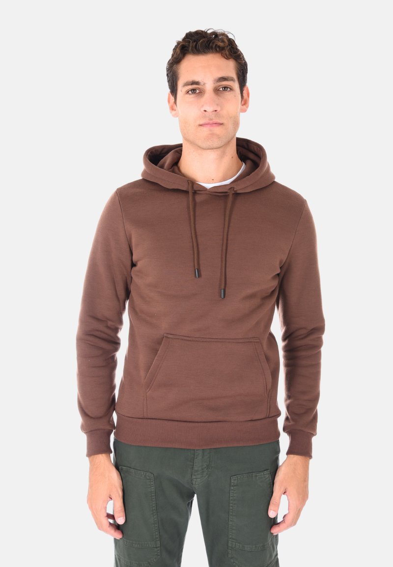 Giovane uomo con capelli corti e ricci, indossa una felpa con cappuccio marrone e pantaloni cargo verde scuro, in piedi su uno sfondo neutro.