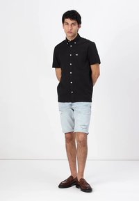 Camisa negra de manga corta con botones y un bolsillo de parche, combinada con shorts de mezclilla claros con detalles desgastados y mocasines marrones.