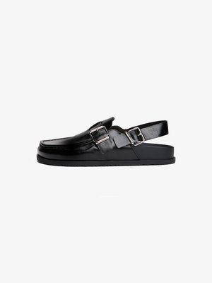Zapato negro de piel tipo slingback con puntera redondeada, que presenta un acento de hebilla y una suela de goma plana. Textura suave, diseño minimalista.