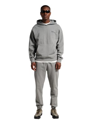 Man draagt een grijze hoodie en bijpassende joggers, beige en zwarte sneakers, en zwarte zonnebril, staand tegen een witte achtergrond.