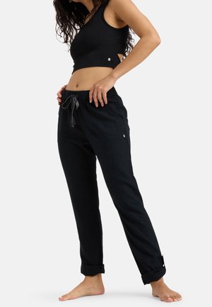 Vrouw draagt een zwart mouwloos crop top en zwarte broek met trekkoord, staat blootsvoets met handen in de zakken tegen een witte achtergrond.
