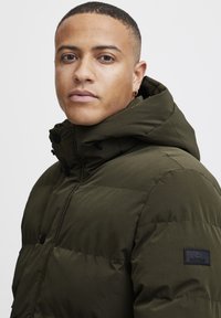 Tumes roheline puhv jacket kapuutsiga, millel on tikitud disain, mustad nupud ja logoga lapp vasakul varrukal. Sile tekstuur.