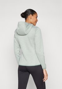 Haut à capuche vert clair avec un motif géométrique, en tissu texturé, à manches longues, et avec un cordon de serrage. Porté avec des leggings noirs.