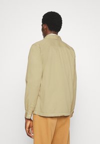 Camisa de manga larga color caqui con cuello, puños abotonados y ajuste holgado. Tela de algodón suave con detalle de yugo en la espalda.