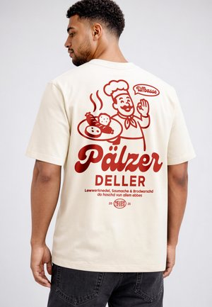 Mann trägt cremefarbenes T-Shirt mit rotem Grafik eines lächelnden Kochs, der einen Teller mit Wurst, Knödeln und Brot hält, mit deutschem Text.