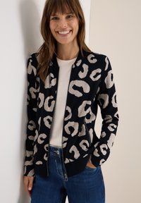 Veste bomber en bleu marine avec imprime léopard beige. Dotée d'une fermeture éclair sur le devant, de poignets et d'un ourlet côtelés, ainsi que de poches latérales. Portée par-dessus un haut clair.