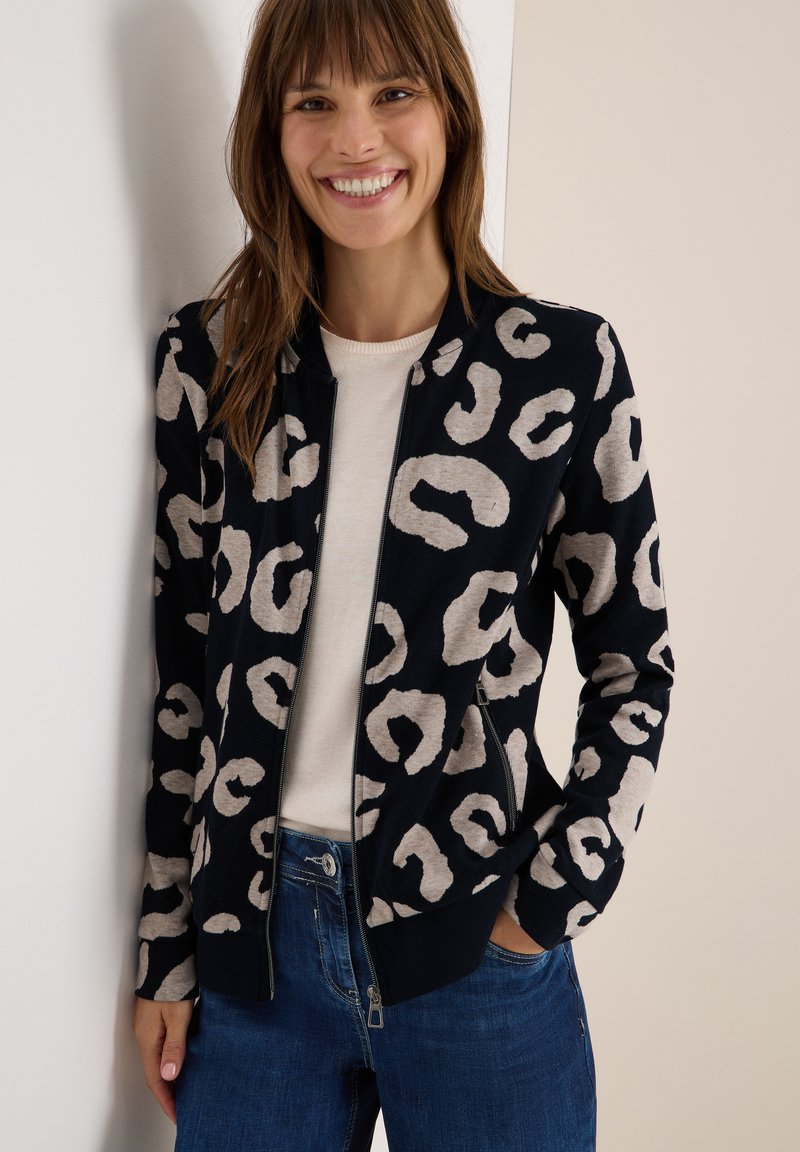 Veste bomber en bleu marine avec imprime léopard beige. Dotée d'une fermeture éclair sur le devant, de poignets et d'un ourlet côtelés, ainsi que de poches latérales. Portée par-dessus un haut clair.