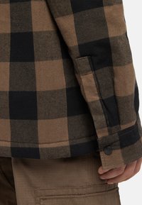 Brun och svart rutigt jacka med knäppning vid ärmslutet, tillverkad av en texturerad ullblandning. Närbild på ärm och hand.