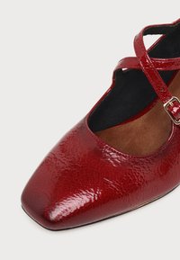Chaussure plate en cuir rouge brillant avec un bout pointu, des brides croisées à la cheville et une petite boucle sur fond clair.